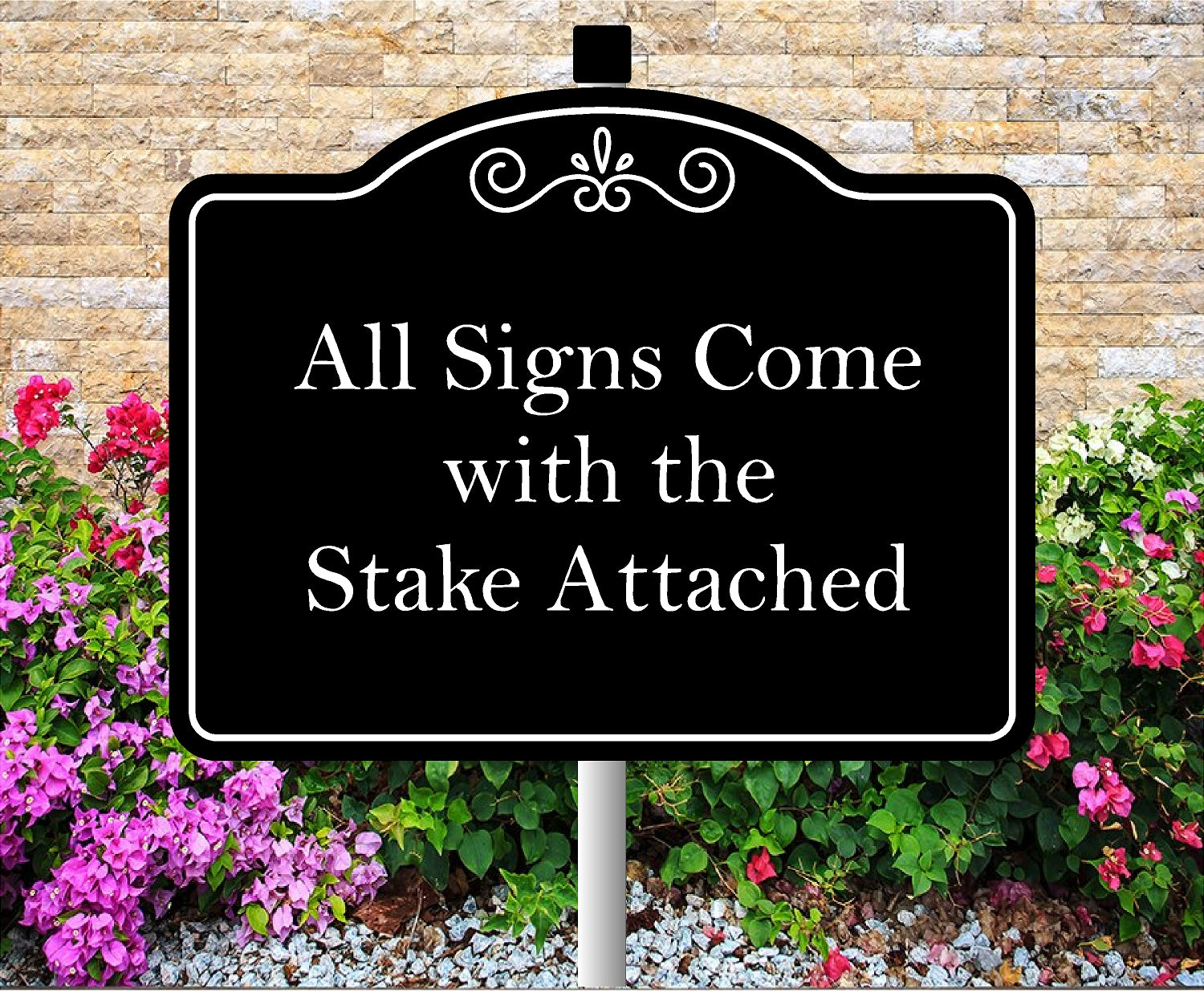 Black sign with white text 'All Signs Come with the Stake Attached' in front of a stone wall and flowers.