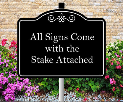Black sign with white text 'All Signs Come with the Stake Attached' in front of a stone wall and flowers.