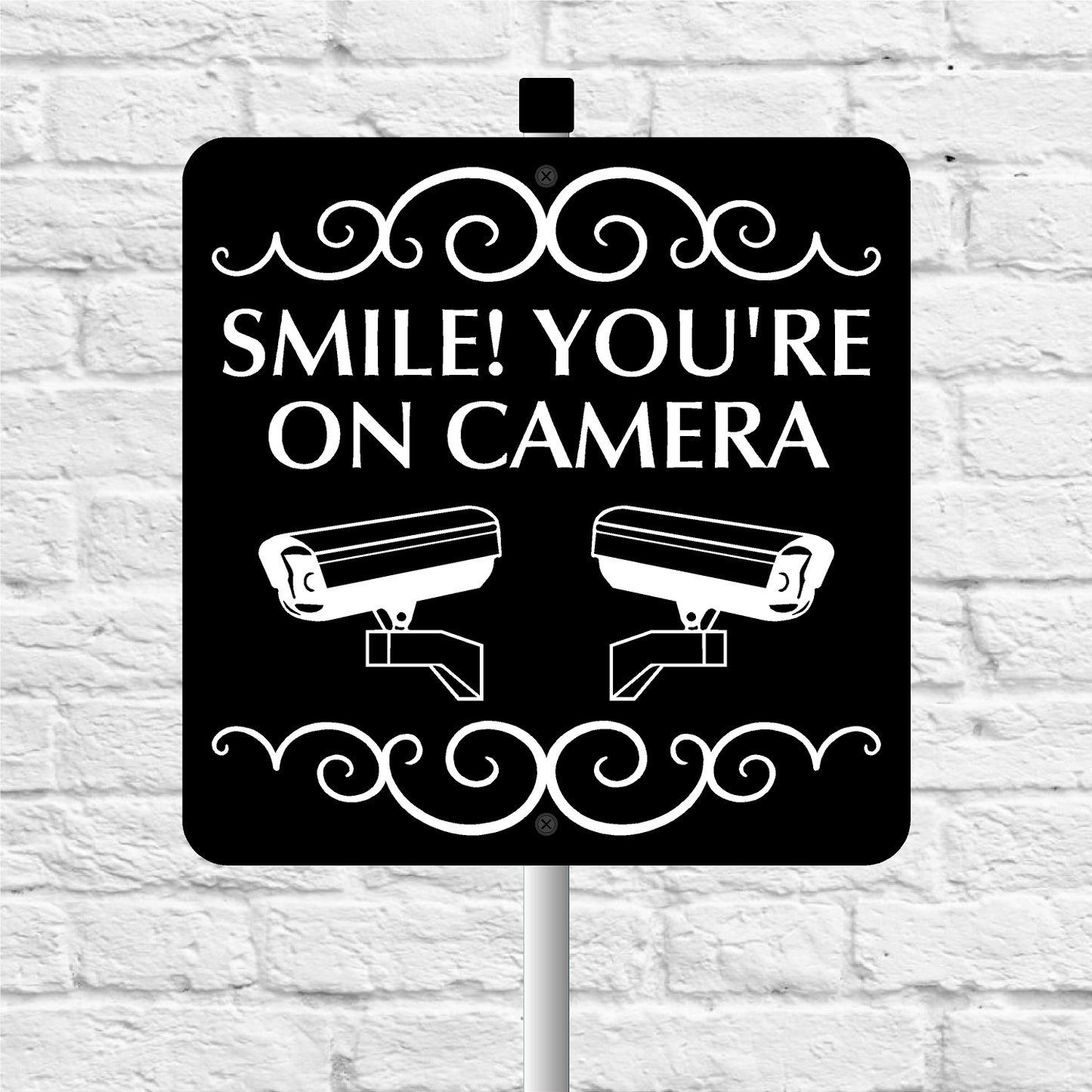 Black sign with 'Smile! You're on camera' text and security camera icons on a white brick wall background.