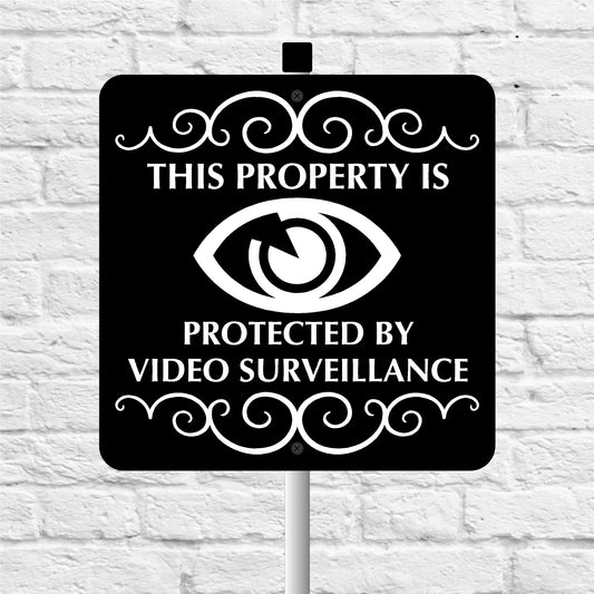Black video surveillance sign and stake with white text and eye symbol on a white brick wall background