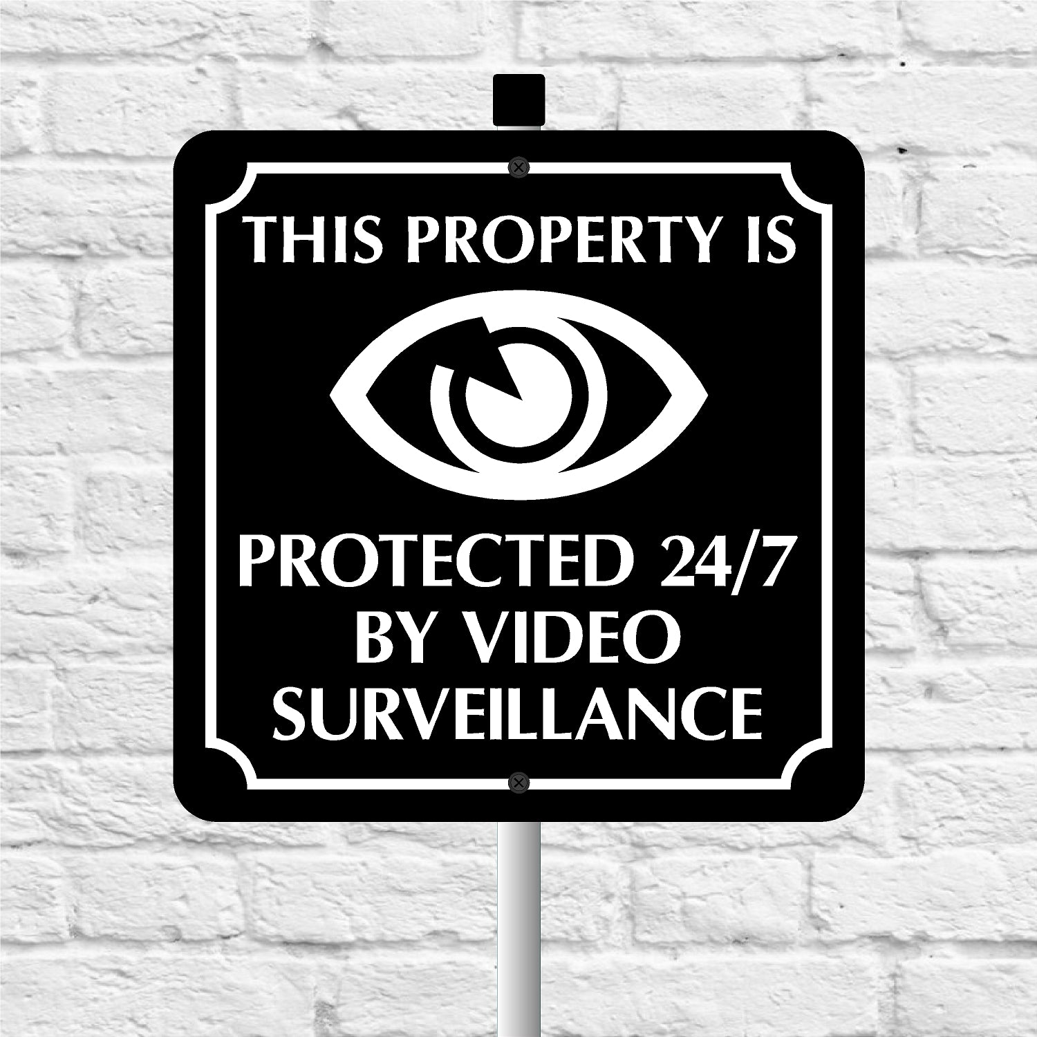 Sign with eye symbol and text 'This property is protected 24/7 by video surveillance' on a white brick wall background.