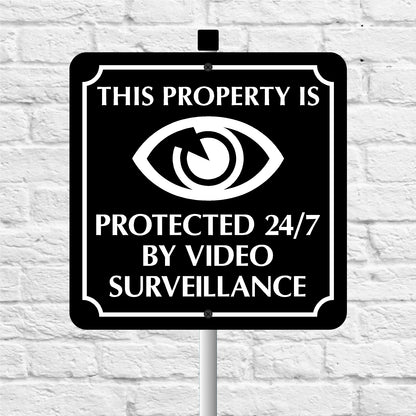 Sign with eye symbol and text 'This property is protected 24/7 by video surveillance' on a white brick wall background.