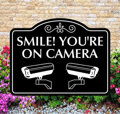 Outdoor security sign with 'Smile! You're on camera' text and camera icons, against a brick wall and flower background.