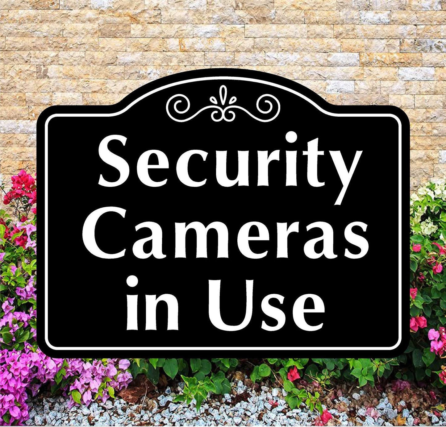 Black sign with 'Security Cameras in Use' text in front of a stone wall and flowers.