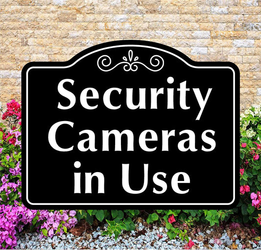 Black sign with 'Security Cameras in Use' text in front of a stone wall and flowers.