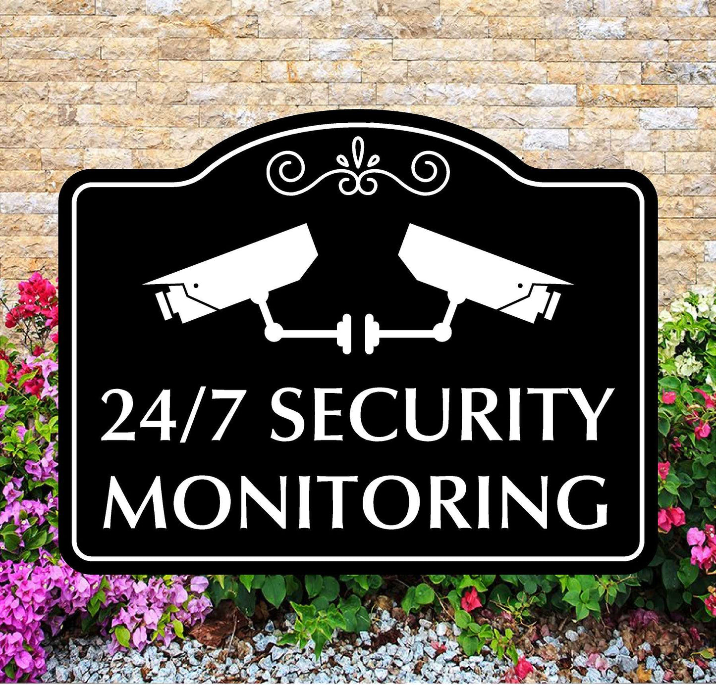Security monitoring sign with two cameras on a black background, surrounded by flowers and gravel.
