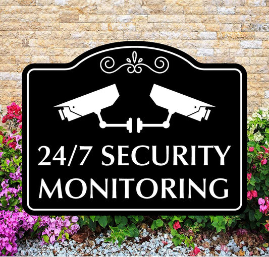 Security monitoring sign with two cameras on a black background, surrounded by flowers and gravel.
